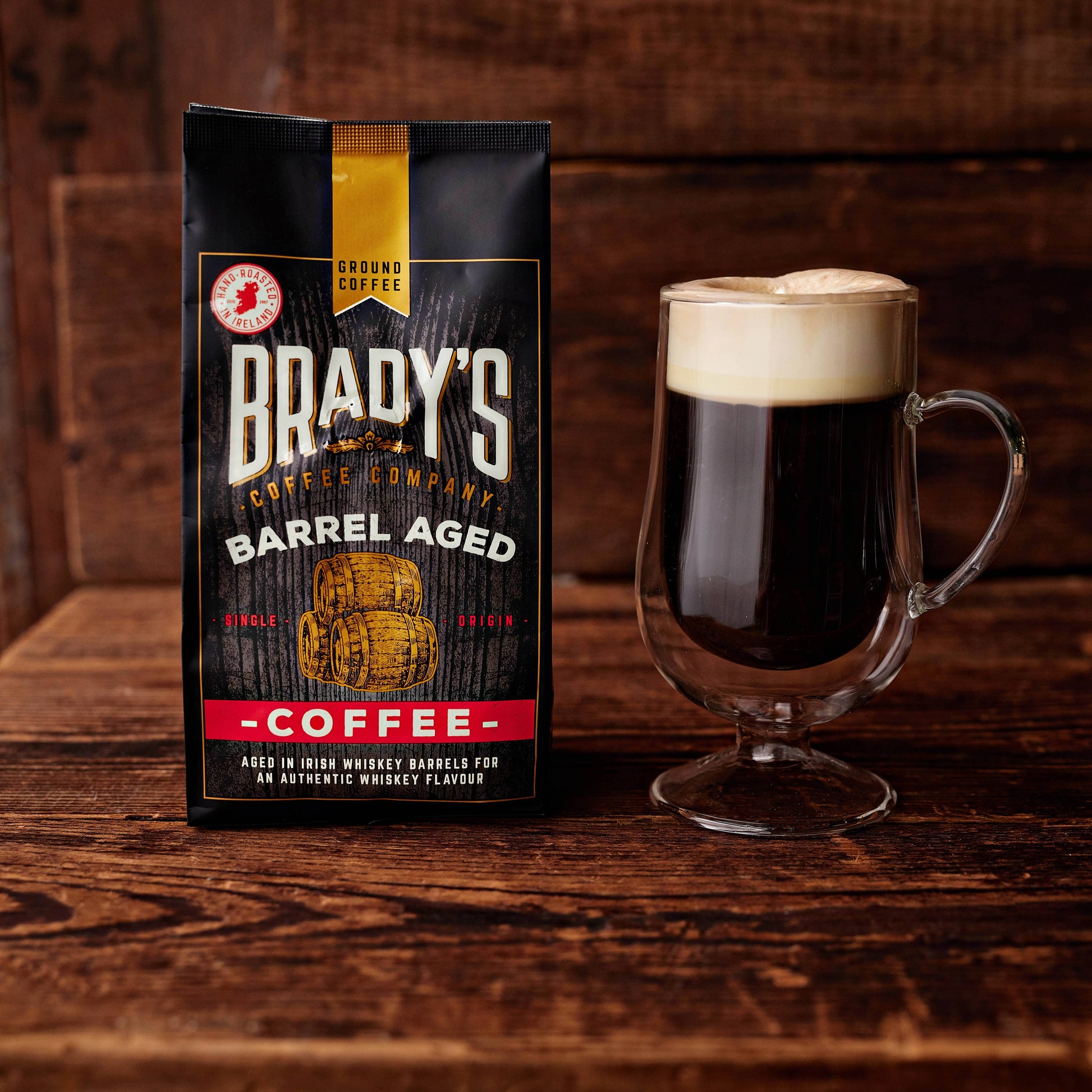 Eine Brady's Coffee Company Kaffeedose mit ganzen, im Fass gereiften Bohnen, irischem Whiskey-Aroma steht neben einer Glas-Tasse auf Holz – ein außergewöhnlicher Genuss für Liebhaber besonderer Aromen.