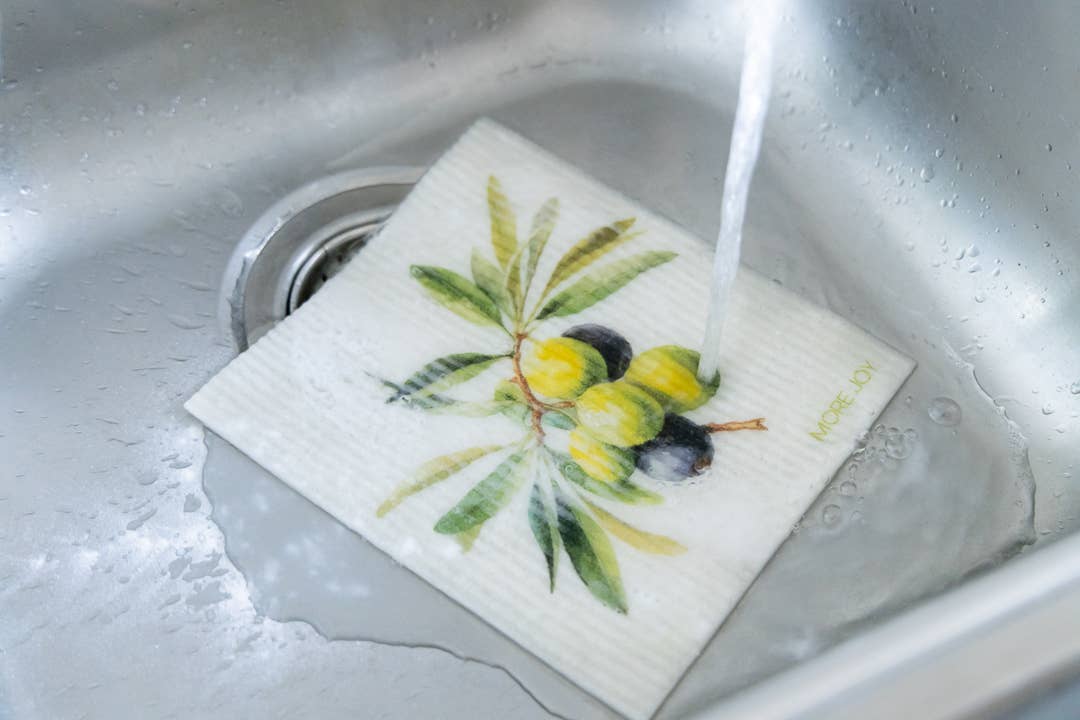 Das More Joy Schwammtuch Olives, ein biologisch abbaubares Schwammtuch im Olivenzweig-Design, liegt flach in der Küchenspüle unter fließendem Wasser aus dem Wasserhahn.