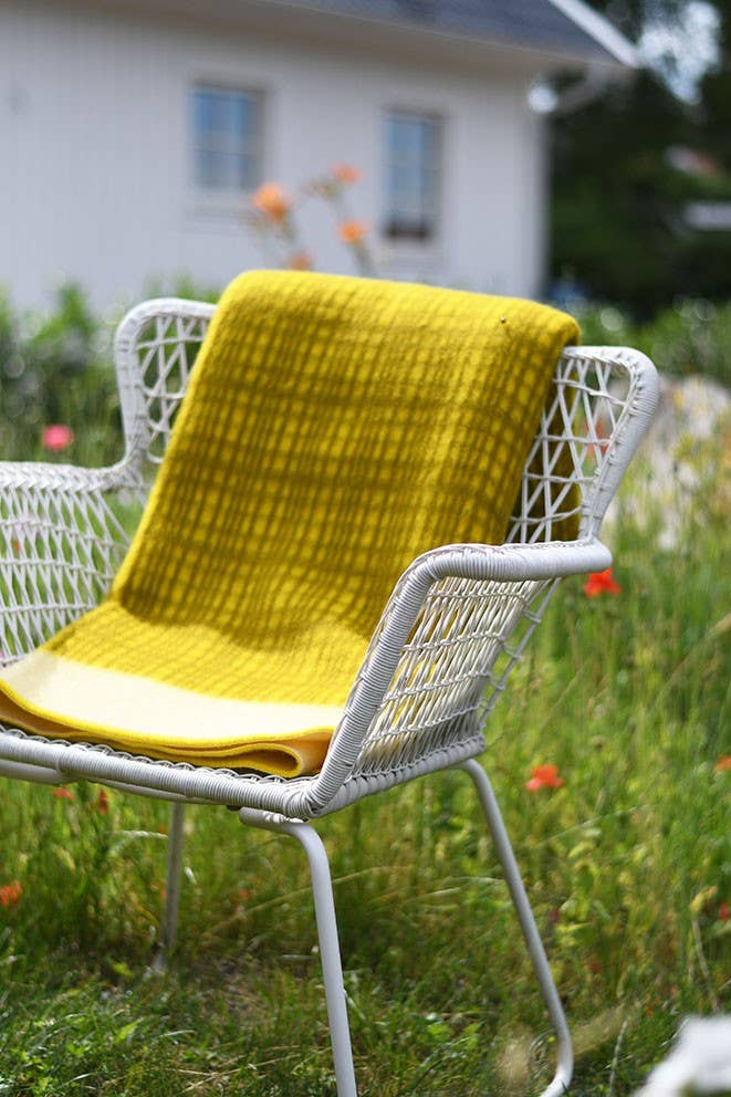 Ein weißer Metallstuhl mit geflochtener Sitzfläche sitzt im Gras, drapiert mit der gelben Darling Tagesdecke aus Lammwolle von Sofie Sjöström Design. Im Hintergrund vervollständigen ein weißes Gebäude und bunte Gartenblumen die Szene.