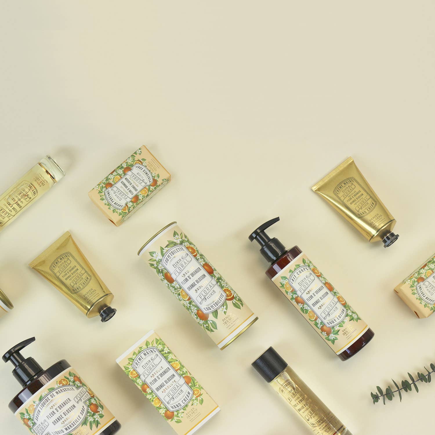 Ein Flat Lay mit Panier Des Sens Duftseife | Orangenblüte 150 g, mit goldener und blumiger Verpackung, Flaschen mit Pumpen, Tuben, natürlichen Inhaltsstoffen und einem Eukalyptuszweig auf einem hellbeigen Hintergrund.