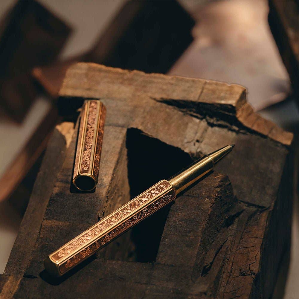 Der ENTREPÔT - YSTUDIO Klassische Renaissance KAZARI KANAGU ORCHID Tintenroller mit seinen kunstvollen Gravuren ruht auf strukturiertem Holz. Die passende Kappe liegt daneben, während warmes Licht die luxuriösen Metalldetails des Stiftes hervorhebt.