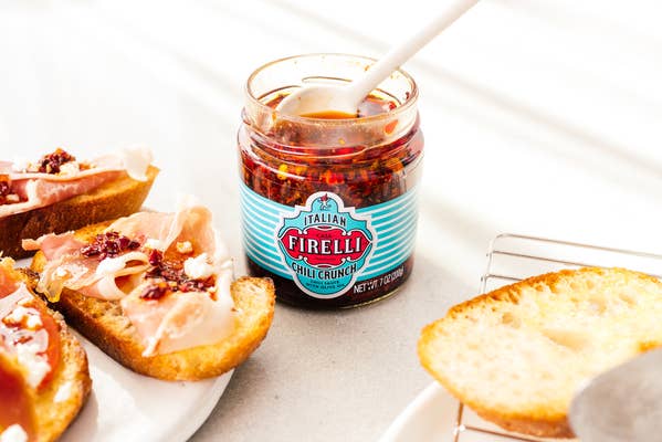 Ein Glas Firelli – Casa Firelli Chili Crunch 200 g Glas steht neben geröstetem Brot mit Chili Crunch und Fleisch, alles auf einer hellen Oberfläche angerichtet.