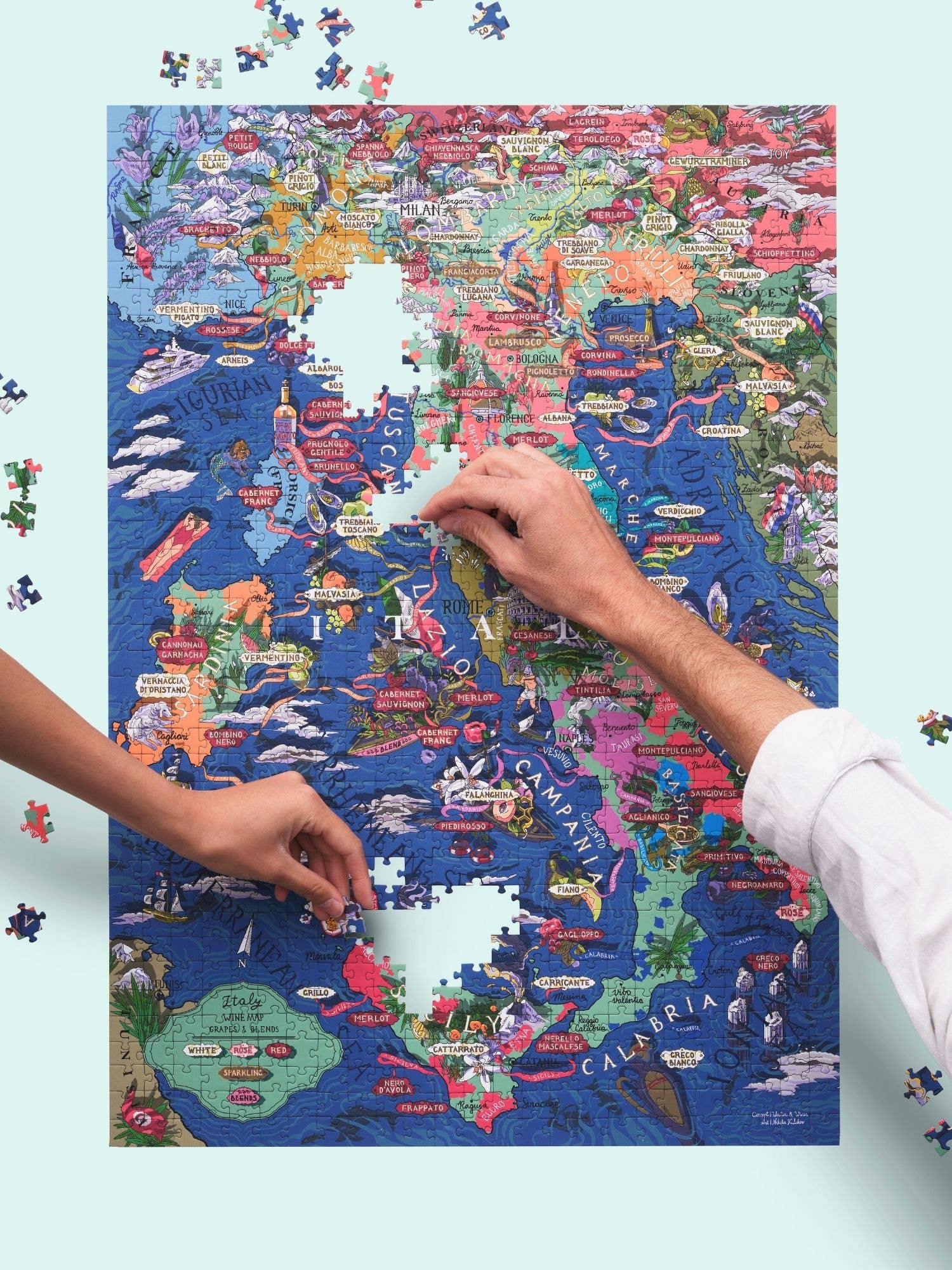 Zwei Personen setzen das Water & Wines - Wein Puzzle - Italien von Water & Wines zusammen, eine farbenfrohe illustrierte Karte mit italienischen Weinregionen und Rebsorten, wobei einige Teile im mittleren und südlichen Teil fehlen.