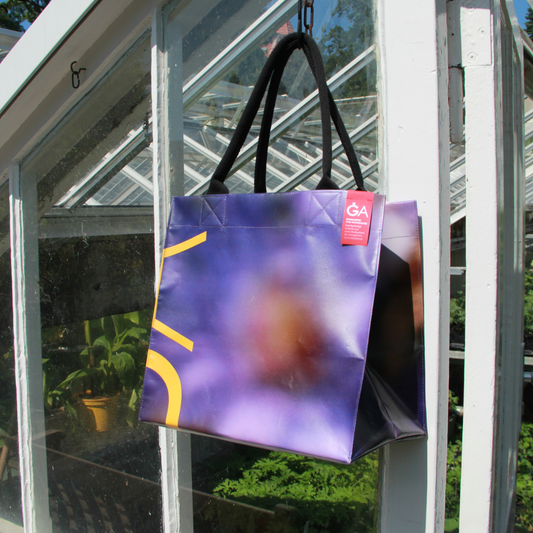 Die Upcycling KGA Tasche 'Aster' von der Königlichen Gartenakademie, eine große wiederverwendbare Tasche mit abstrakten lila und gelben Motiven, hängt an einer weißen Gewächshaustür mit Sonnenlicht und Grünzeug im Hintergrund.