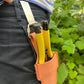 Eine Person in schwarzen Hosen hat einen Niwaki-Gürtelclip an ihrem Hosenbund befestigt und hält zwei Werkzeuge mit gelben Griffen, im Hintergrund ist grünes Laub zu sehen.