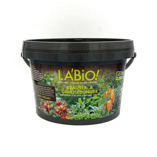 Ein schwarzer Plastikbecher mit der Aufschrift La'Bio Kräuter- & Gemüsedünger von La'Bio, auf dem Bilder von Tomaten und Kräutern zu sehen sind, weist darauf hin, dass es sich um einen veganen Dünger für eine umweltfreundliche Ernährung von Kräutern und Gemüse handelt.