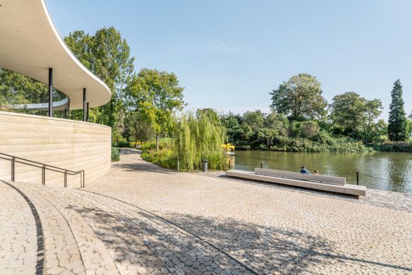 Ein modernes, geschwungenes Gebäude mit hellen Steinwänden blickt auf eine gepflasterte Terrasse an einem ruhigen Fluss, umgeben von Grün - eine inspirierende Szene, die in "Landschaftsarchitektur 2025" aus dem Callwey Verlag vorgestellt wird. Bänke säumen das Wasser; zwei Personen entspannen sich in der Nähe.