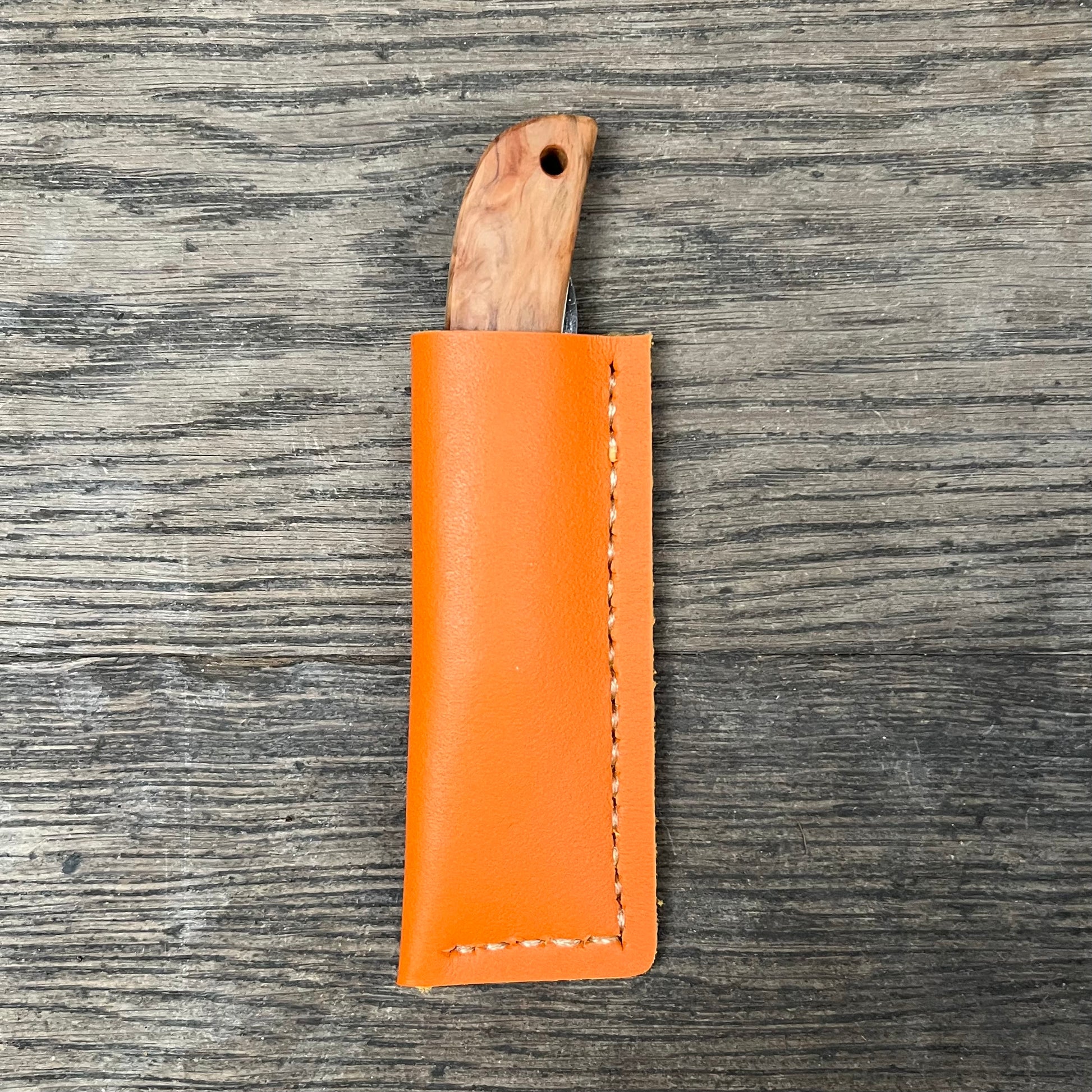 Ein Niwaki Kotoh X Niwaki-Messer mit Holzgriff liegt teilweise in einer orangefarbenen Lederhülle, wobei sich die leuchtende Scheide vom strukturierten Holz abhebt – von Niwaki für höchste Schneidleistung gefertigt.