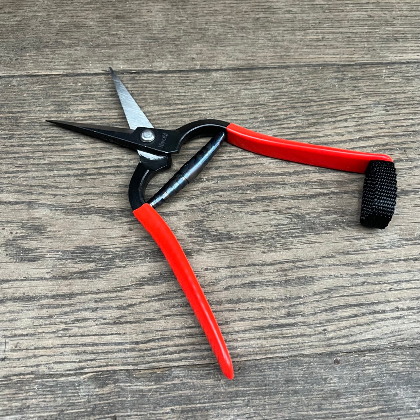 Eine Niwaki Forged Snips von Niwaki mit roten Griffen und einer schwarzen Feder liegt offen auf einer Holzoberfläche.