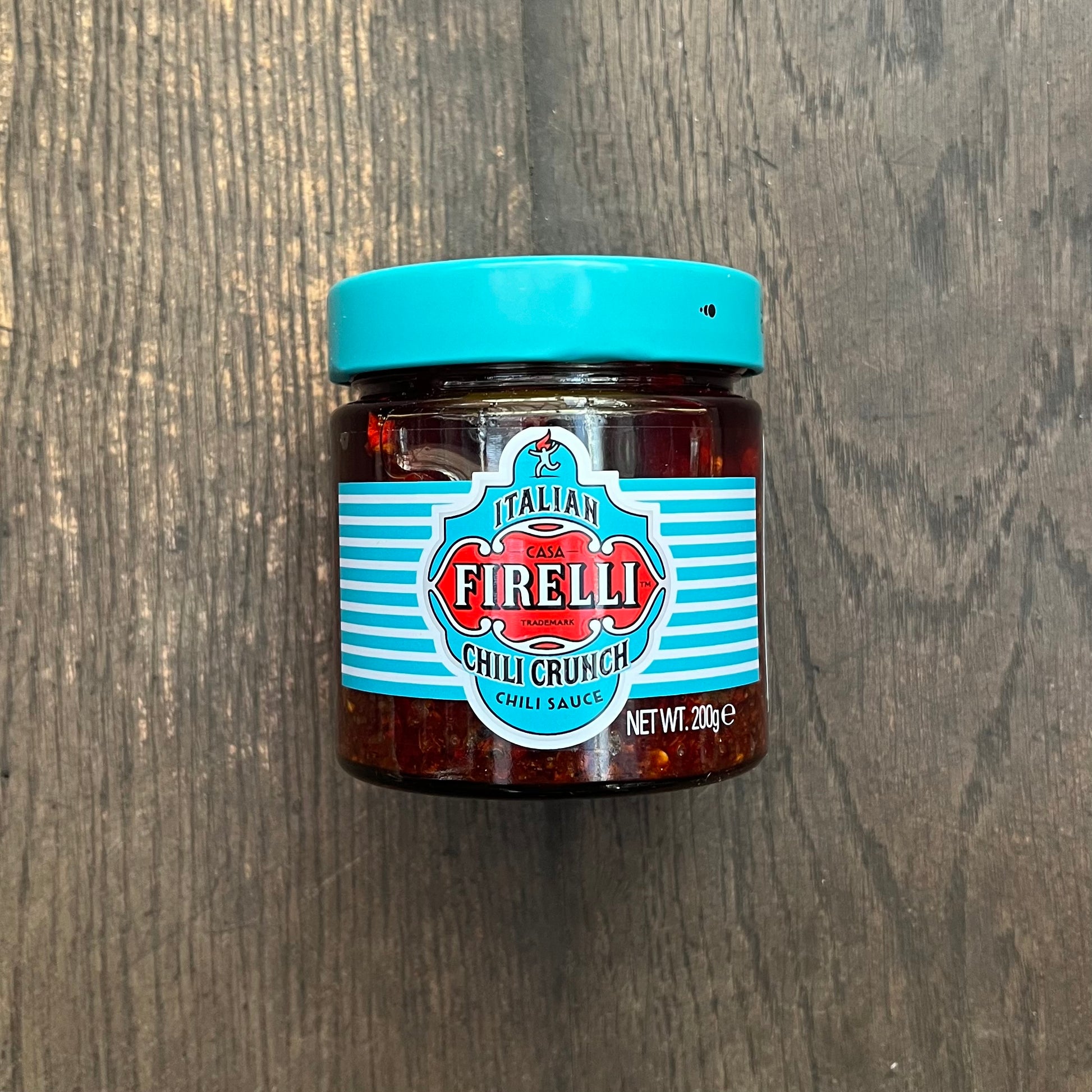 Ein Glas Firelli - Casa Firelli Chili Crunch 200g Glas von Firelli mit türkisfarbenem Deckel und Etikett steht auf einer dunklen Holzoberfläche. Hergestellt aus italienischem Olivenöl und kalabrischen Chiliflocken, ist das Etikett mit rot-weißem Text und dekorativen Streifen versehen.