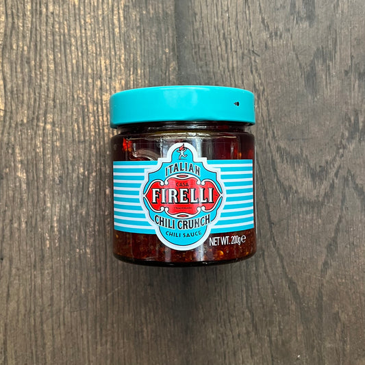Ein Glas Firelli - Casa Firelli Chili Crunch 200g Glas von Firelli mit türkisfarbenem Deckel und Etikett steht auf einer dunklen Holzoberfläche. Hergestellt aus italienischem Olivenöl und kalabrischen Chiliflocken, ist das Etikett mit rot-weißem Text und dekorativen Streifen versehen.