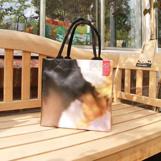 Die Upcycling KGA Tasche 'Torf' von Königliche Gartenakademie mit schwarzen Henkeln und einzigartigem Werbebanner-Print steht samt rotem Etikett auf einer Holzbank im Grünen vor großen Glasfenstern.