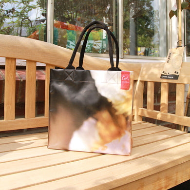 Die Upcycling KGA Tasche 'Torf' von Königliche Gartenakademie mit schwarzen Henkeln und einzigartigem Werbebanner-Print steht samt rotem Etikett auf einer Holzbank im Grünen vor großen Glasfenstern.