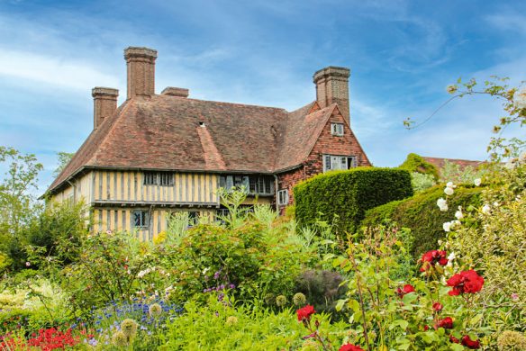 Hinter einem üppigen, farbenfrohen Garten, der von "Gärtnern wie Gott in England" aus dem Callwey Verlag inspiriert wurde, steht ein traditionelles englisches Cottage mit rotem Ziegeldach und hohen Schornsteinen unter einem strahlend blauen Himmel, umgeben von blühenden Blumen und Grünzeug.