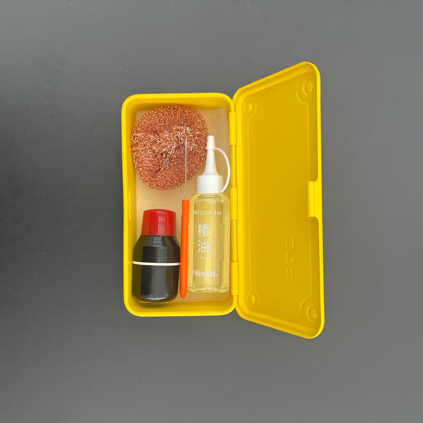 Ein offener gelber Niwaki T-Type-Werkzeugkasten enthält eine Ölflasche, einen schwarz-roten Behälter und einen Kupferschrubber, alles angeordnet auf einem hellbraunen Schaumstoffeinsatz – perfekt für Gärtner und Handwerker.