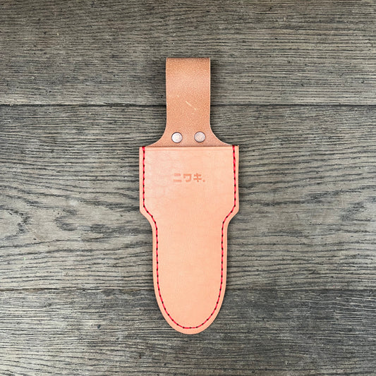 Ein hellbraunes Niwaki-Einzelholster von Niwaki mit roten Nähten und zwei Metallnieten liegt auf einer Holzoberfläche. Auf der Vorderseite eingeprägte japanische Schriftzeichen verleihen Ihrer Schere Stil und schützen sie gleichzeitig.