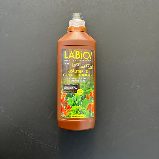Eine Flasche La'Bio Kräuter & Gemüsedünger von LaBio, ein 100% veganer Dünger mit grünem und gelbem Etikett, auf dem Tomaten und Kräuter abgebildet sind, steigert den Pflanzenertrag und ist vor einem schwarzen Hintergrund abgebildet.