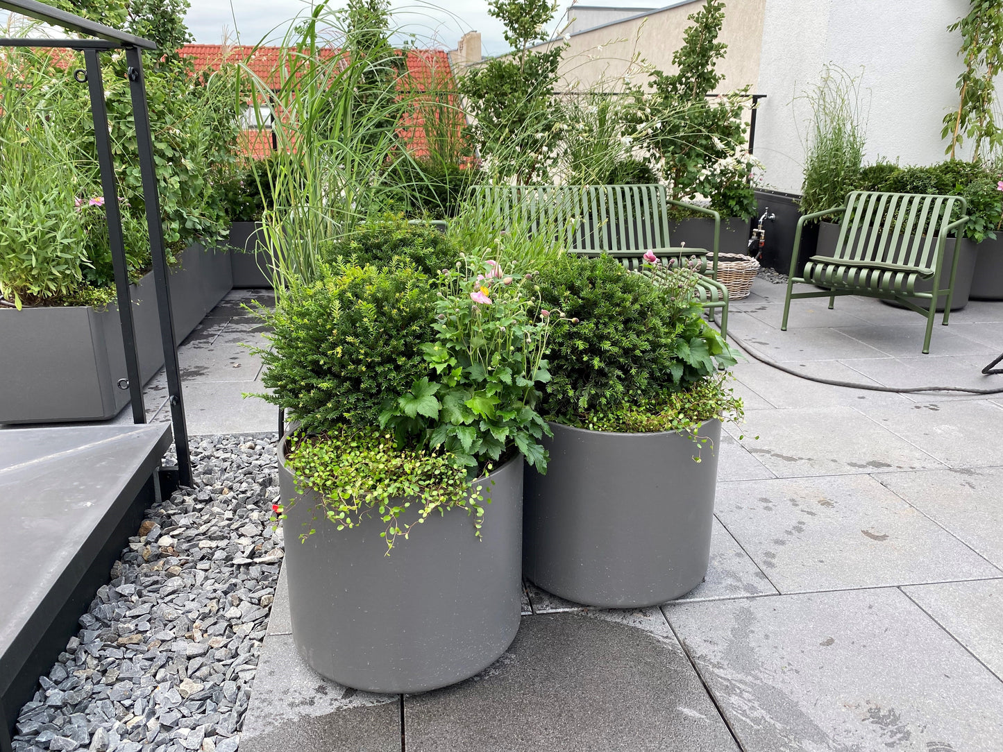 Zwei große graue Pflanzgefäße mit nachhaltigen Pflanzen begrünten eine moderne Dachterrasse, die in der Ausstellung "Di. 10.03.2026 | 10.00-13.00 HALBTAGESKURS Dachterrassen und Balkonbepflanzung" von Astrid Torrens, umgeben von grünen Stühlen und Schotter.