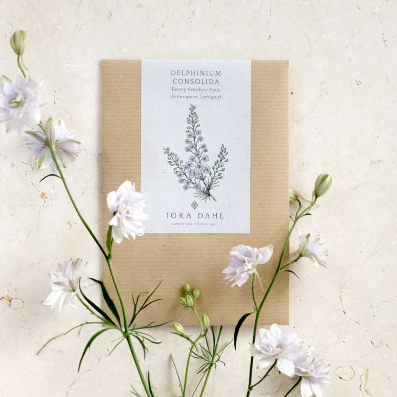 Ein braunes Saatgutpaket mit der Aufschrift Jora Dahl | Delphinium consolida 'Fancy Smokey Eyes' (Rittersporn), mit einer botanischen Illustration - ideal für einjährige Blumen oder Schnittblumen - inmitten hellvioletter Blüten auf einem strukturierten Hintergrund.