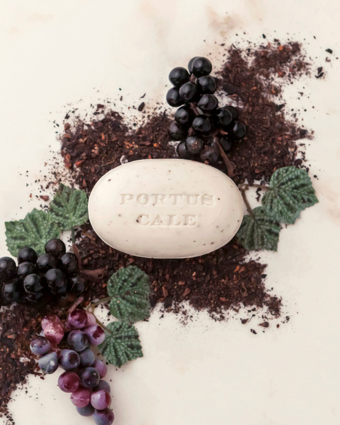 A Castelbel Seife Portus Cale 150 g - Rubinrot liegt auf weißem Untergrund, gerahmt von Teeblättern, künstlichen Trauben und grünen Blättern - eine pflanzliche Duftseife mit Trauben-Beeren-Aroma.