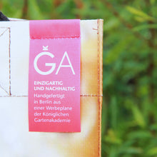 Lade das Bild in den Galerie-Viewer, Nahaufnahme eines Stoffanhängers mit deutschem Text auf der Upcycling KGA Tasche &quot;Torf&quot; der Königlichen Gartenakademie, der besagt, dass es sich um ein Unikat handelt, das in Berlin aus einem einmaligen Werbebanner handgefertigt wurde, vor einem unscharfen grünen Hintergrund.
