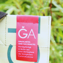 Lade das Bild in den Galerie-Viewer, Eine Nahaufnahme eines Unikat-Taschen-Etiketts mit deutschem Text, das auf dem cremefarbenen Upcycling KGA Shopper &#39;Orange&#39; der Königlichen Gartenakademie angebracht ist. Unscharfes grünes Laub erscheint im Hintergrund.
