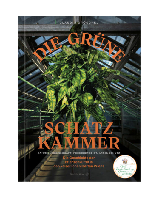 Das Buchcover von Die grüne Schatzkammer aus dem Brandstätter Verlag zeigt einen leuchtend grünen Farn in einem gläsernen Gewächshaus im Schlosspark Schönbrunn, mit fettgedruckter gelber Schrift und einem runden Preisaufkleber in der rechten unteren Ecke.