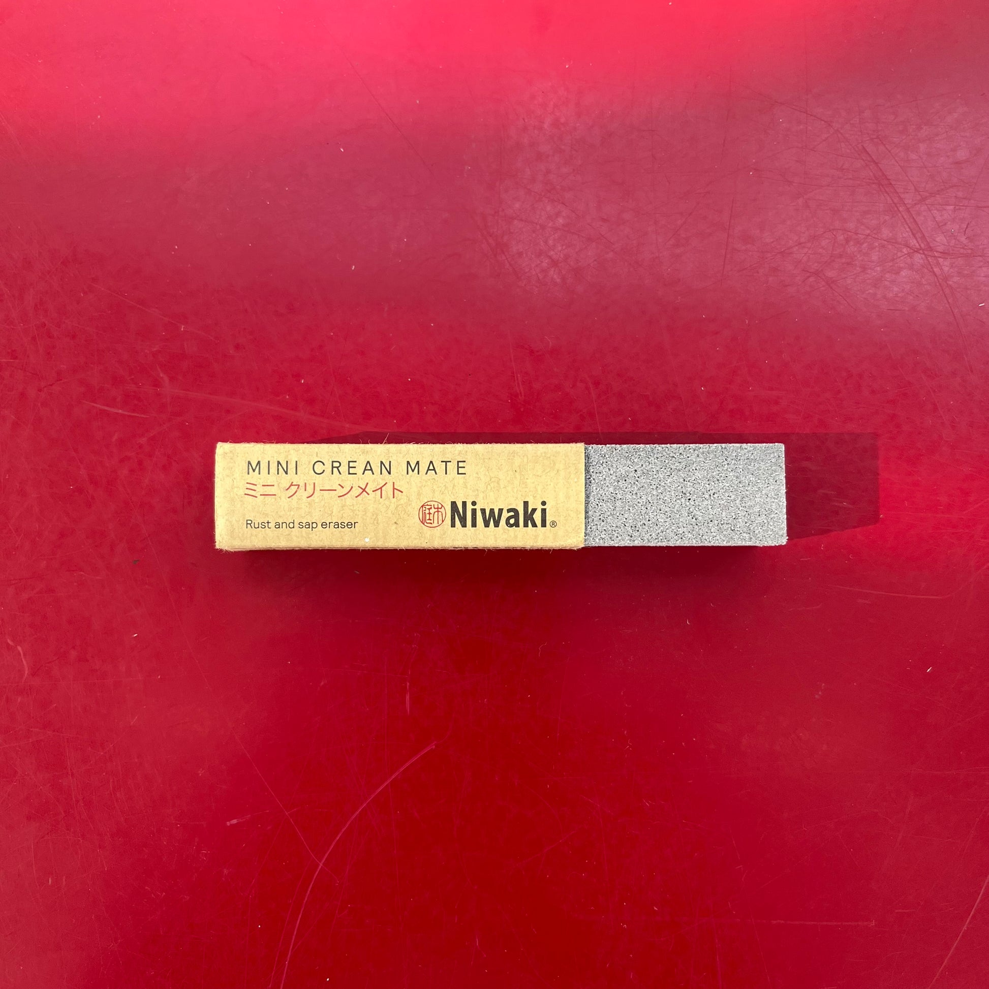 Ein Niwaki - Mini Crean Mate Rost- und Saftradierer mit beigem Etikett und grauem Radiergummi ruht auf einer roten Oberfläche. Ideal für die Reinigung Gartenscheren, sein Etikett verfügt über Englisch und Japanisch Text. Marke: Niwaki.
