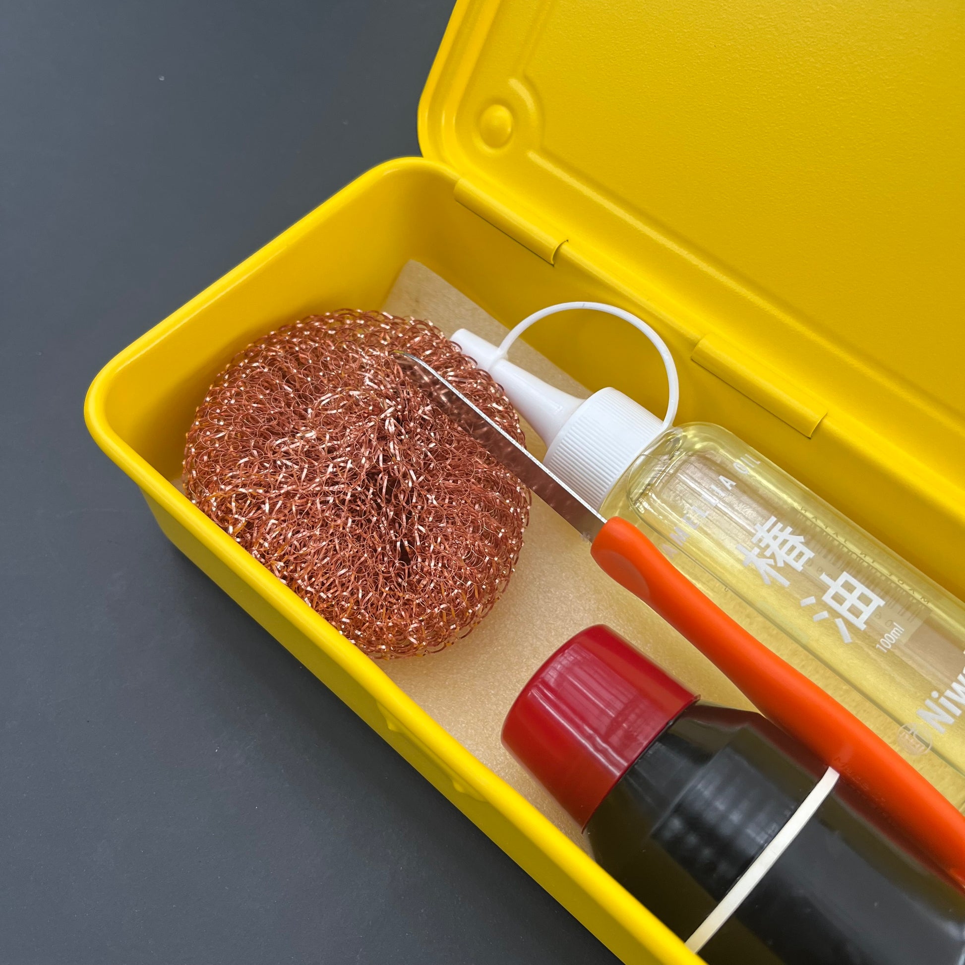 Eine Niwaki T-Type Toolbox von Niwaki in Gelb enthält einen Kupferschrubber, eine durchsichtige Sprühflasche und eine schwarze Flasche mit rotem Deckel, alles ordentlich angeordnet auf einer dunklen Oberfläche.
