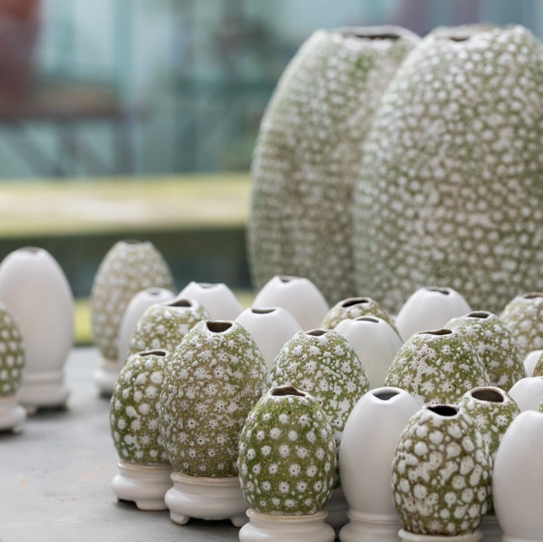 Ein Satz kleiner "Vasen von Des pots | Ei-auf-Schale Form Klein" der Königlichen Gartenakademie, einige schlicht weiß und andere mit grün-weißen Blumenmustern, schmückt den Tisch, während zwei größere gemusterte Vasen im Hintergrund stehen.