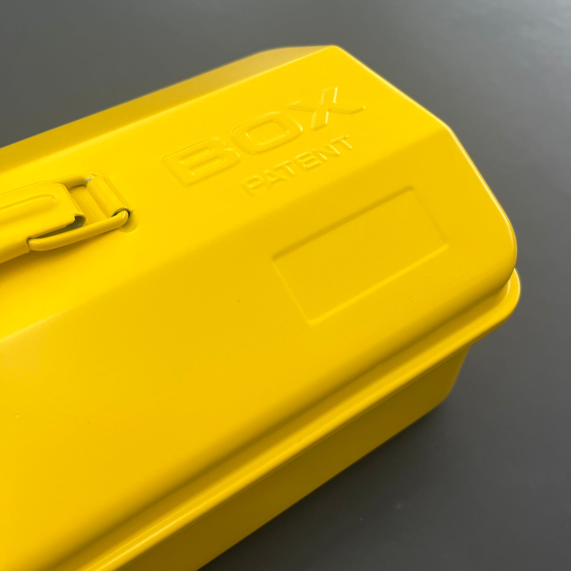 Eine Nahaufnahme einer leuchtend gelben Werkzeugbox Gelb mit "BOX PATENT" auf dem Deckel; links ist ein Metallverschluss zu sehen. Vor einem dunkelgrauen Hintergrund ist sie perfekt für die Aufbewahrung von Gartenutensilien oder Werkzeugen wie die Niwaki Y-Type Toolbox von Niwaki.
