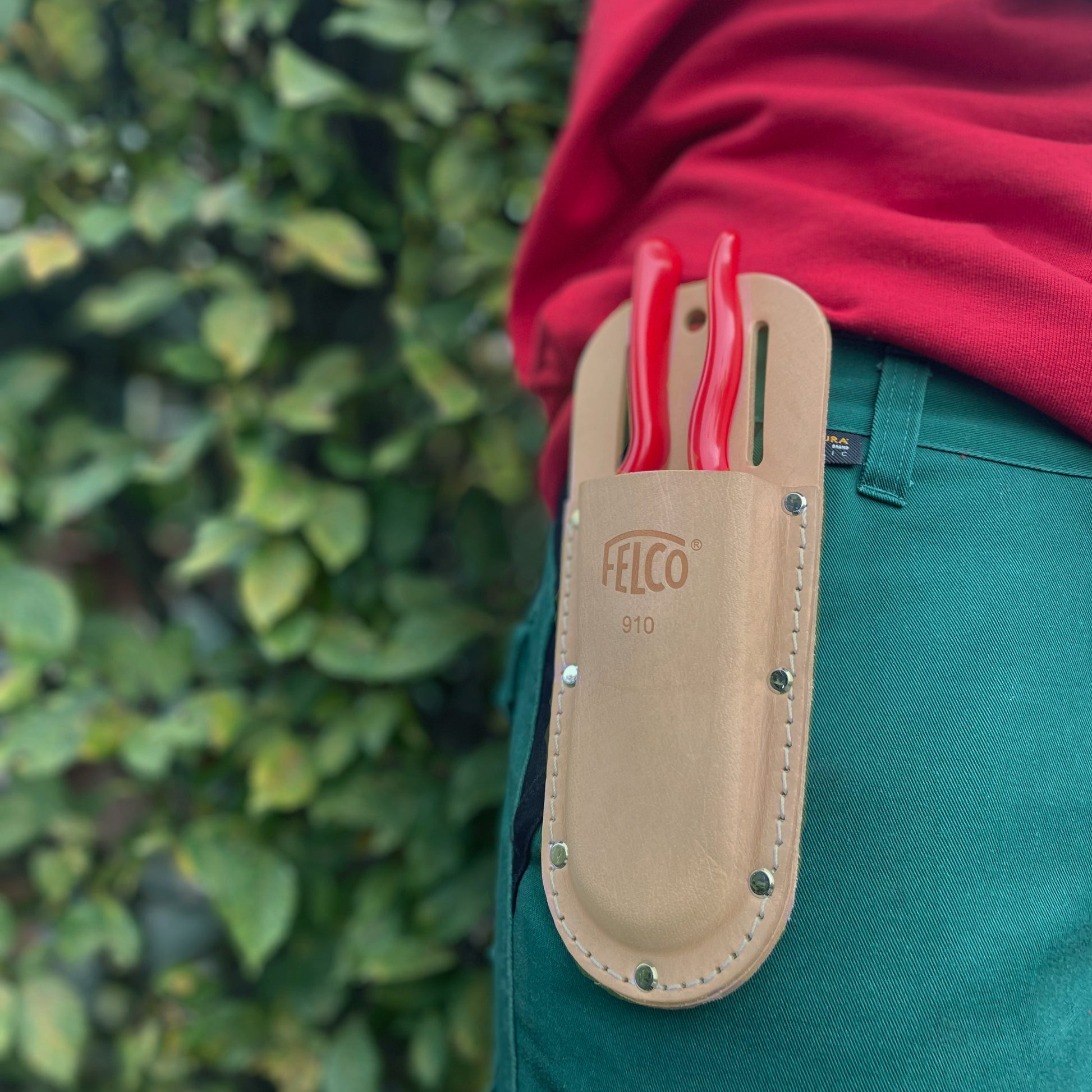 Eine Person in grünen Hosen und einem roten Hemd trägt eine Gartenschere mit rotem Griff in einem hellbraunen Felco 910 Echtleder Messeretui von Felco am Gürtel, im Hintergrund ist grünes Laub zu sehen.