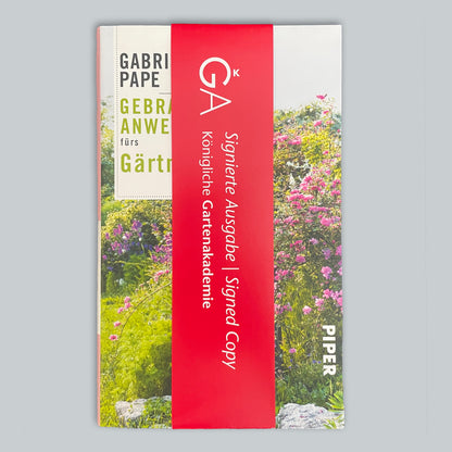 Das Bild zeigt "Buch - Gebrauchsanweisung fürs Gärtnern" von Gabriella Pape, eingewickelt in ein rotes Band mit der Aufschrift "Signierte Ausgabe | Signed Copy, Königliche Gartenakademie". Der Einband zeigt eine farbenfrohe Gartenszene.