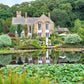 Ein Steinhaus mit gewölbten Fenstern steht inmitten von Gärten, die von "Gärtnern wie Gott in England" aus dem Callwey Verlag inspiriert sind, und spiegelt sich in einem Seerosenteich.