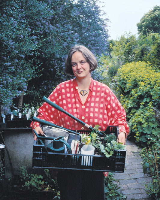 Eine Frau in einem rot karierten Hemd steht mit einer Kiste voller Pflanzen im Freien und versprüht den Charme eines Cottage Gartens - die perfekte Inspiration für Dr. Isabelle Van Groeningens "Sa. 07.03.2026 | 13.30-16.30 HALBTAGESKURS Der Cottage Garten: Der Garten der Zukunft".