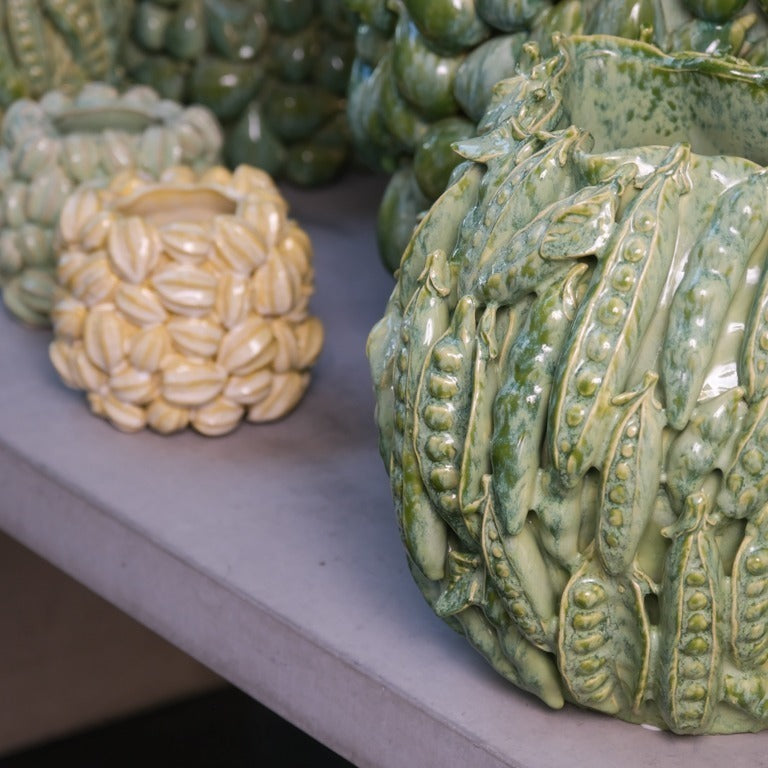 Zwei Keramikvasen von Des Pots stehen auf dem Tisch: Die Vase | Grün mit Schoten ist grün glasiert und mit erhabenen Erbsenschoten verziert, daneben eine kleinere cremefarbene Vase mit ovalen Samendesigns.