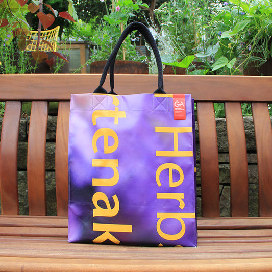 Der Upcycling KGA Shopper 'Herbs' von Königliche Gartenakademie ist eine nachhaltige Unikat-Tasche mit schwarzem Henkel, kleinem roten Anhänger und gelbem Aufdruck auf lila Grund - dekorativ platziert im Grünen.