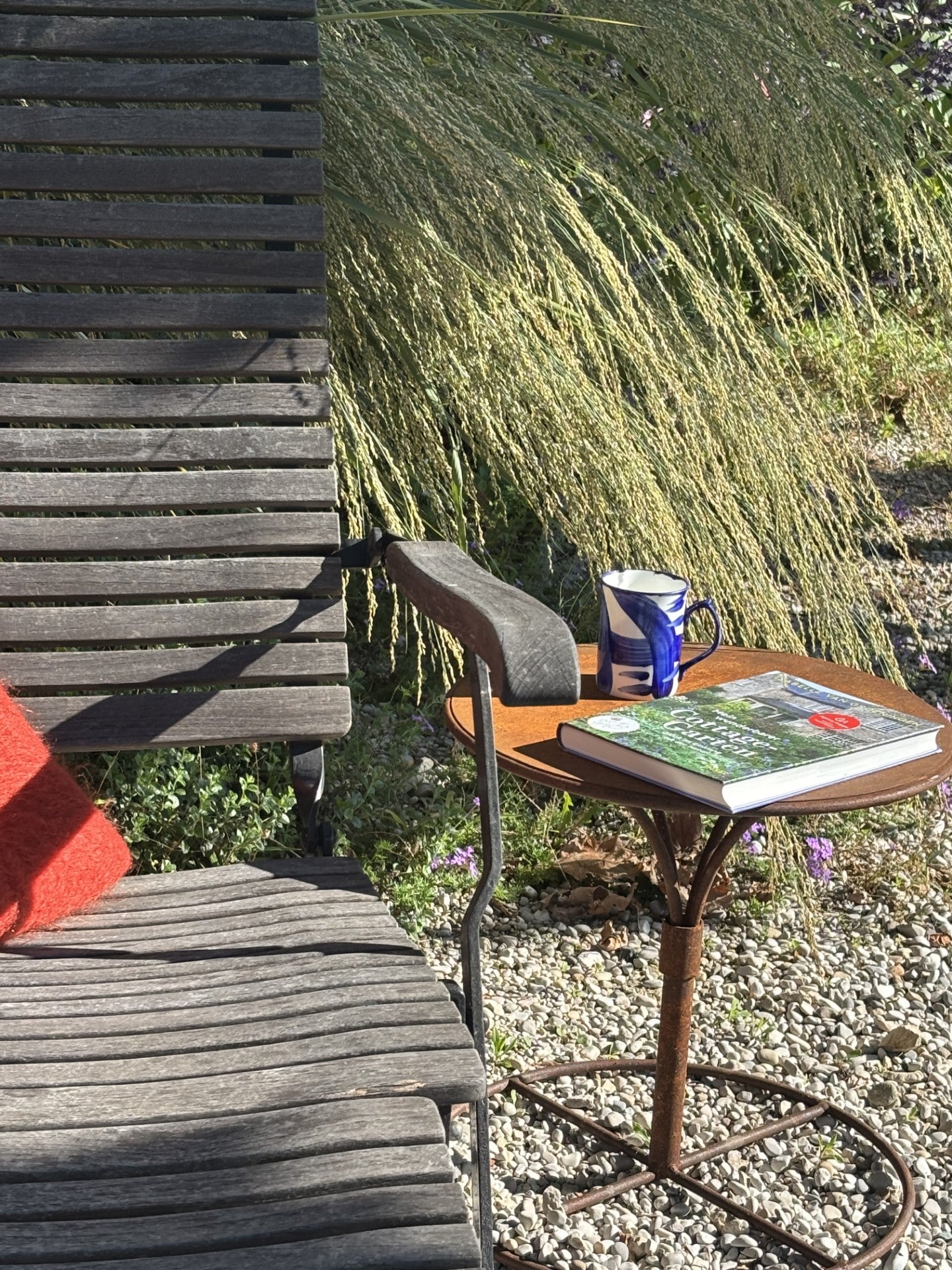 Ein Holzstuhl und ein kleiner Tisch mit einer blau-weißen Tasse und einem geschlossenen Buch stehen inmitten von Kies und hohen Gräsern - eine einladende Kulisse für Gabriella Papes "Mi. 18.02.2026 | 09.30-12.30 HALBTAGESKURS Der Garten als Lebensraum NEU.