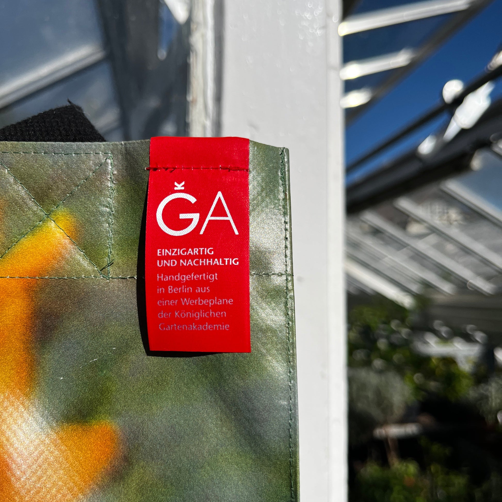 Eine Nahaufnahme der KGA Upcycling Tasche "Gewächshaus" der Königlichen Gartenakademie, handgefertigt in Berlin aus einem wiederverwendeten Gewächshausbanner, zeigt das farbenfrohe Design und das rote "GA"-Tag; im Hintergrund sind Glasplatten zu sehen.
