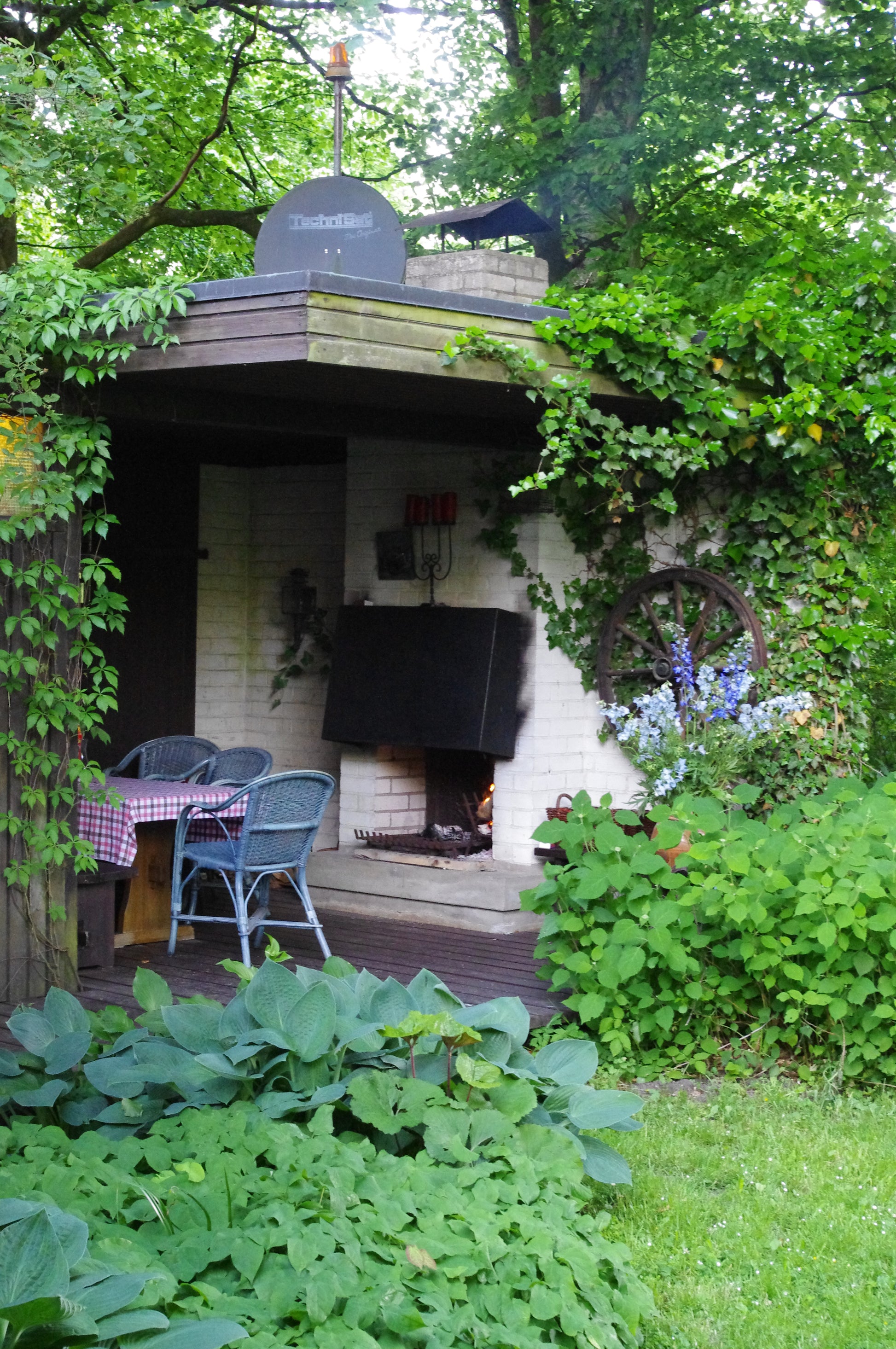 Gestalten Sie eine gemütliche Außenterrasse, inspiriert von Gabriella Papes "Mi. 18.02.2026 | 09.30-12.30 HALBTAGESKURS Der Garten als Lebensraum NEU", mit einem gemauerten Kamin, blauen Korbstühlen, einer Holzbank, einer rot-karierten Tischdecke und üppigen Pflanzen mit Kletterpflanzen.