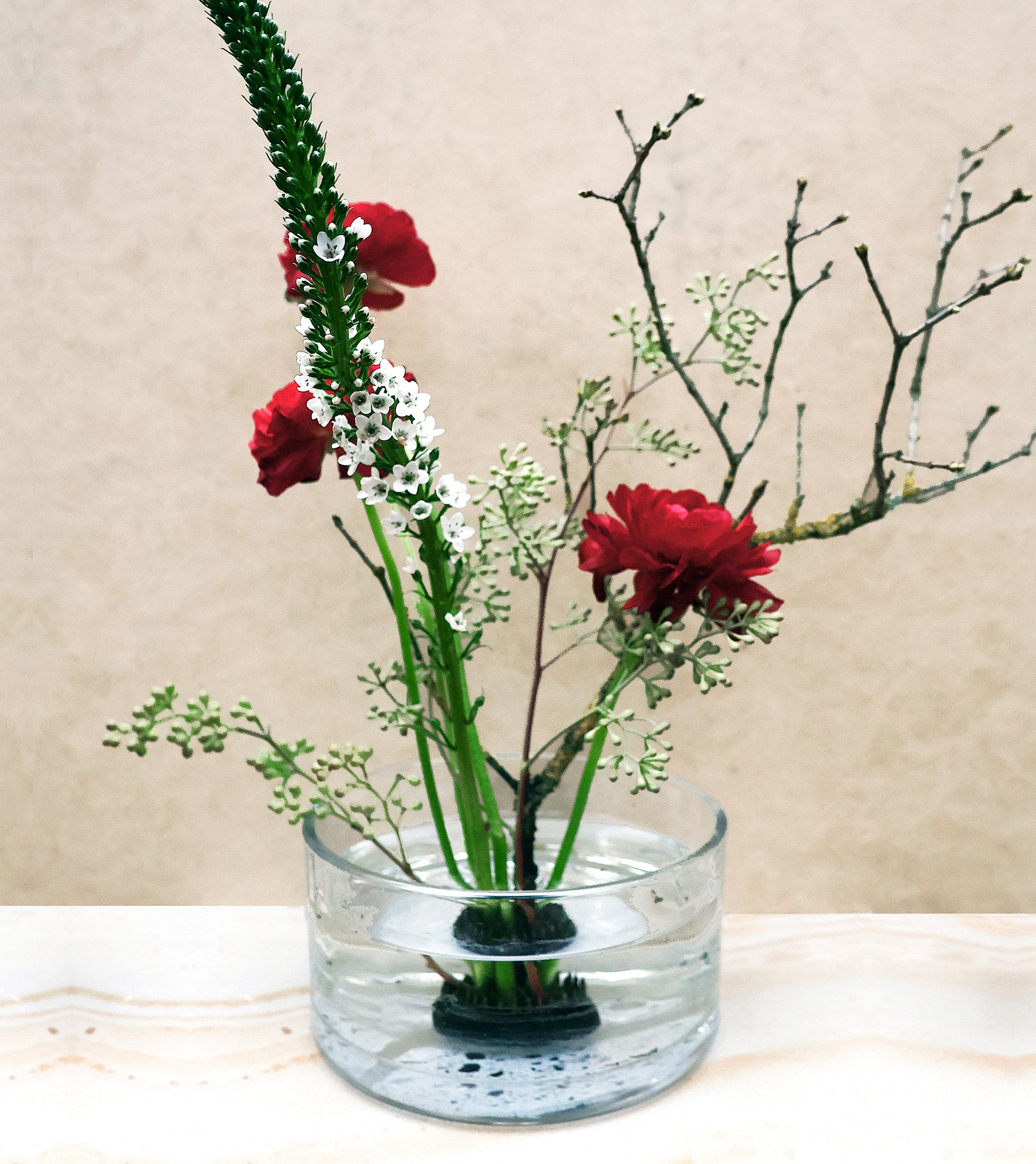 Der Ikebana Blumenigel | Kenzan L zeigt ein minimalistisches Blumenarrangement aus roten Blumen, weißen Blüten, grünen Stängeln und kahlen Zweigen in klarem Wasser vor einem neutralen Hintergrund.