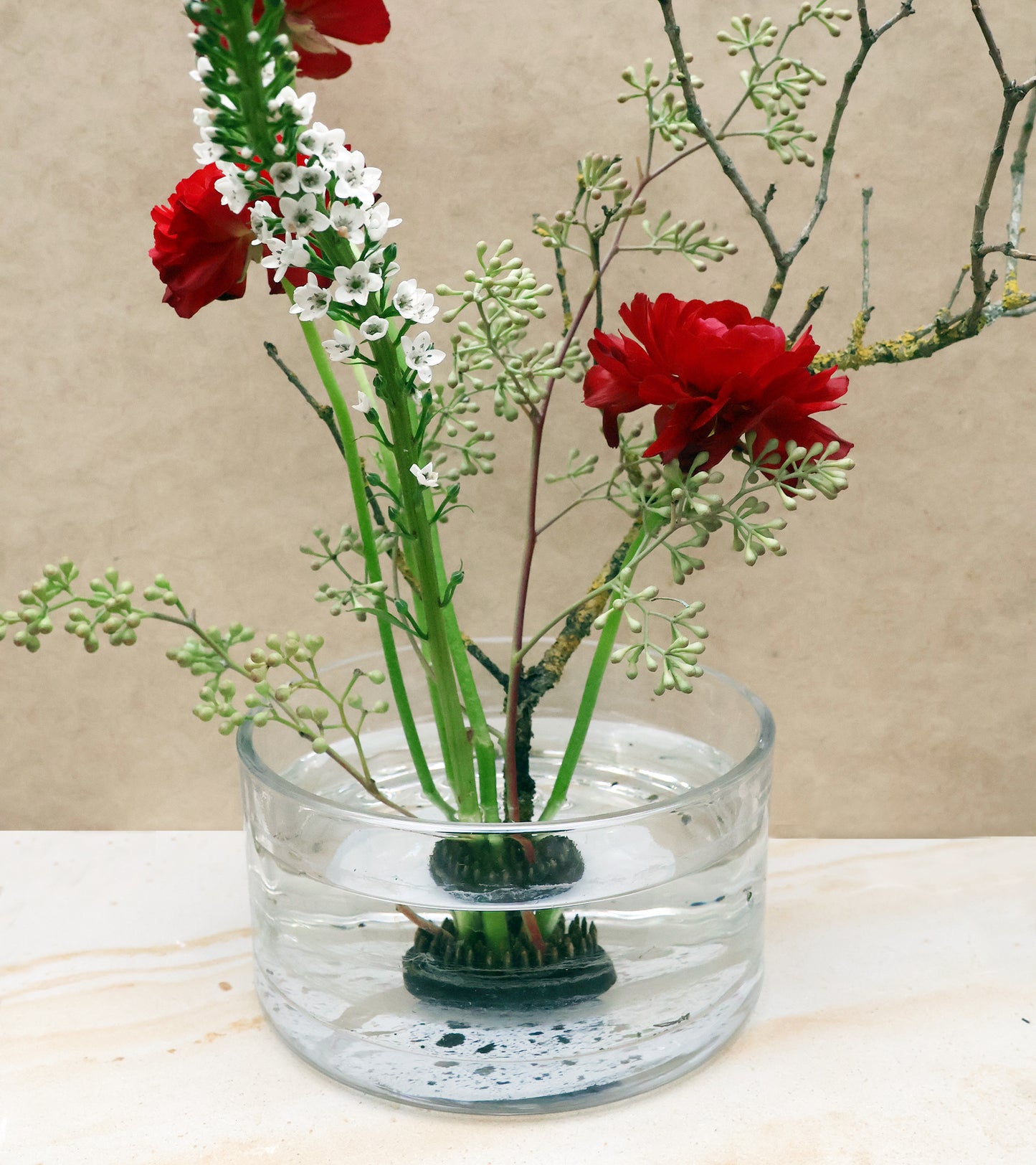 In einer klaren Glasschale mit Wasser stehen rote Blumen, weiße Blüten und grünes Laub aufrecht auf dem Ikebana Blumenigel | Kenzan L Blumenfrosch, arrangiert im klassischen Ikebana-Stil vor einem neutralen Hintergrund.