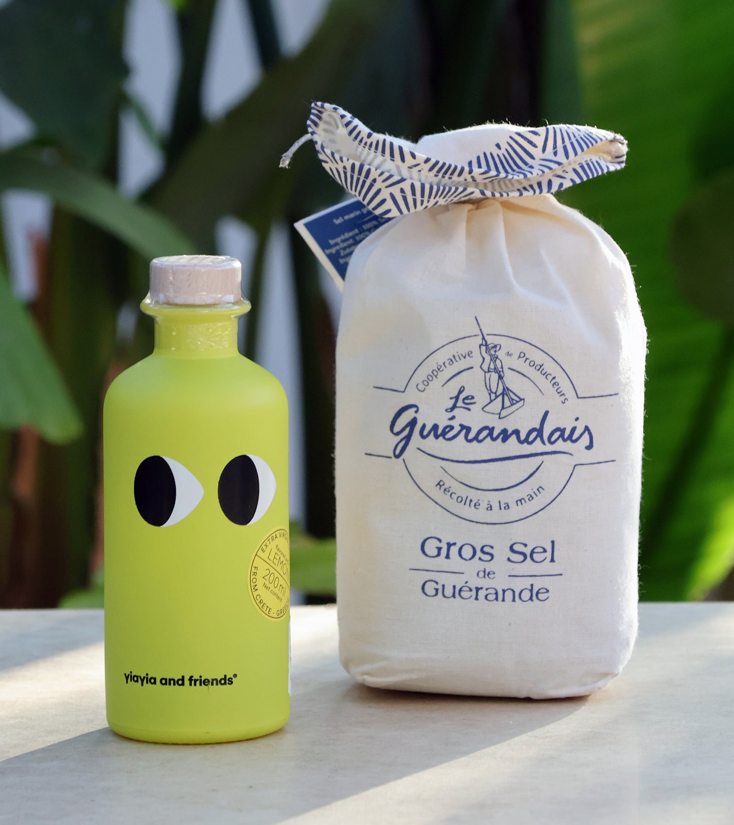 Eine gelbe Yaiya and friends-Flasche mit großen schwarz-weißen Augen steht neben einem Stoffbeutel mit Le Guérandais Gros Sel de Guérande, umgeben von leuchtend grünen Blättern.