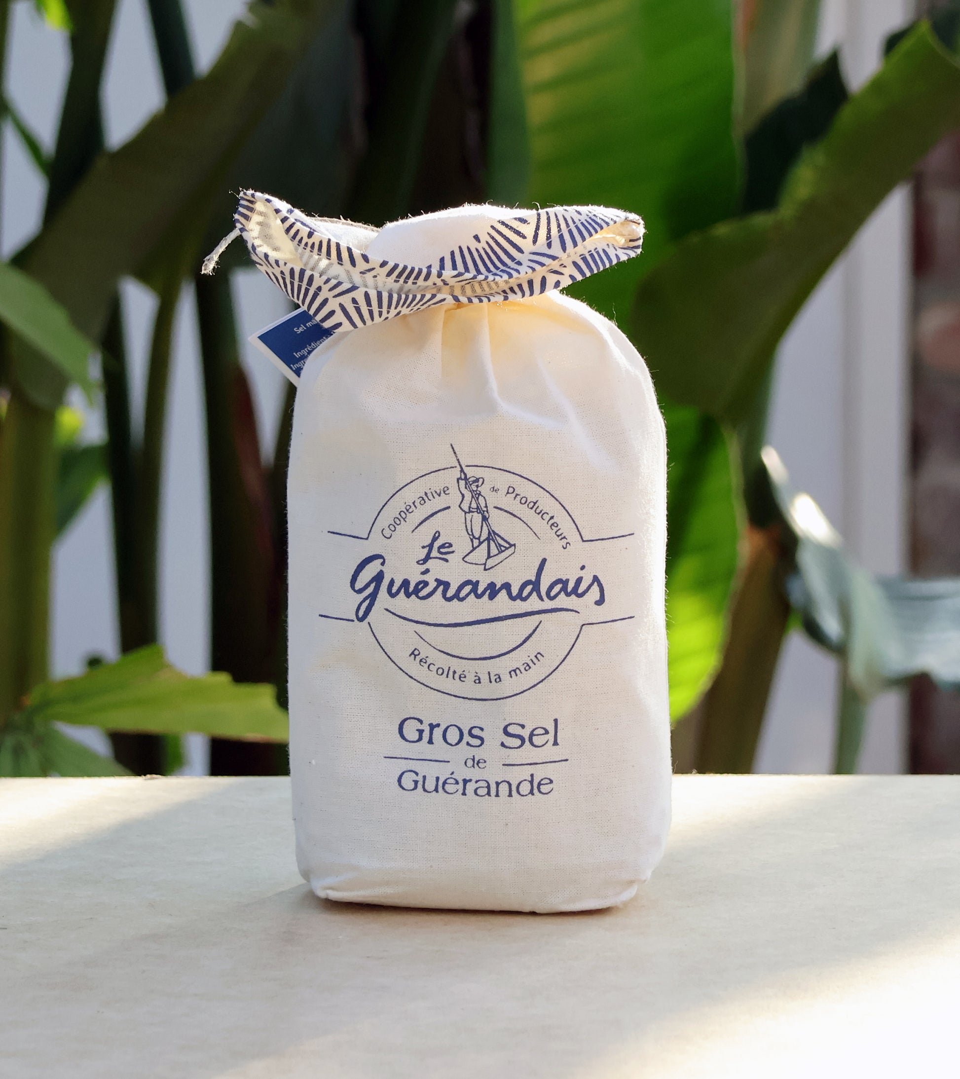 Ein weißer Stoffbeutel mit Meersalz Le Guérandais Guerandais mit blauer Aufschrift und Kordelzug liegt auf einem Tisch im Freien zwischen Grünpflanzen im gedämpften Sonnenlicht.