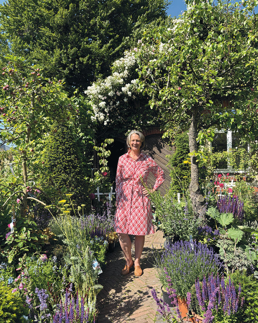 Eine Frau im rot-weißen Kleid lächelt auf einem Gartenweg - eine perfekte Szene für alle, die Gärten als Lebensräume schätzen. Lassen Sie sich inspirieren beim HALBTAGESKURS "Der Garten als Lebensraum" von Gabriella Pape am Mi. 18.02.2026, 09:30-12:30.
