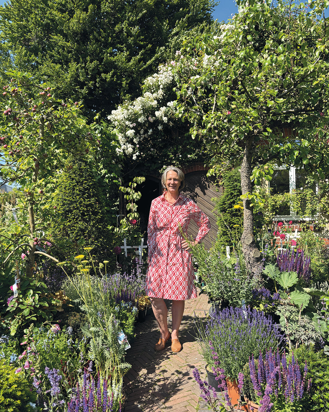 Eine Frau lächelt auf einem Gartenweg inmitten von Blüten und fängt damit den Geist von "Der Garten als Lebensraum NEU" ein - dem Halbtageskurs von Gabriella Pape am Mi. 18.02.2026 von 09:30-12:30 Uhr, perfekt zum Genießen von Gartenräumen gestalten in der Sonne.