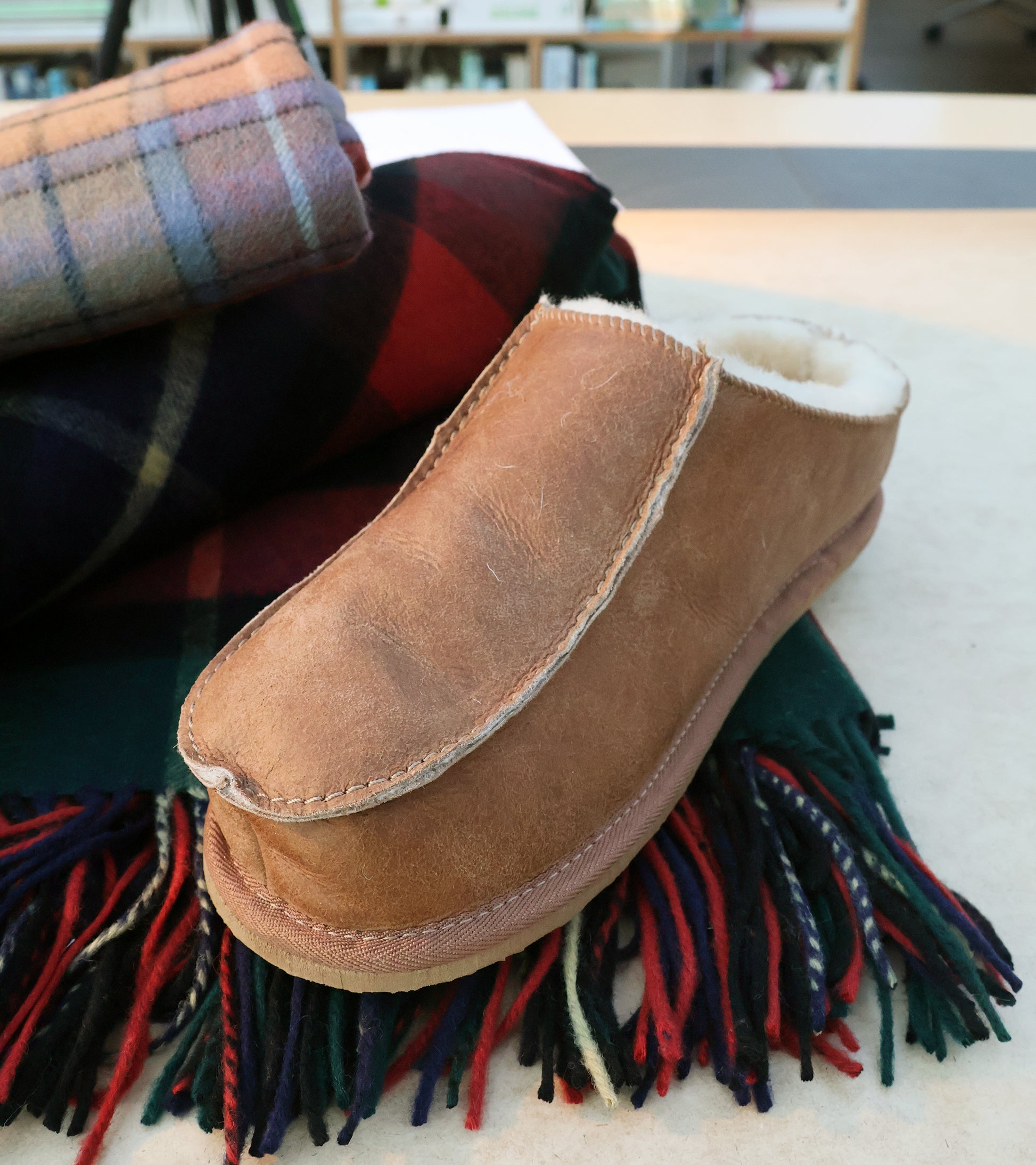 Ein Richard Slipper aus Schaffell in Cognac (Größe 42) von Shepherd of Sweden liegt gemütlich auf einem gewollten Schal mit roten, grünen und marineblauen Streifen; im Hintergrund befindet sich eine zusammengerollte Tartan-Decke.