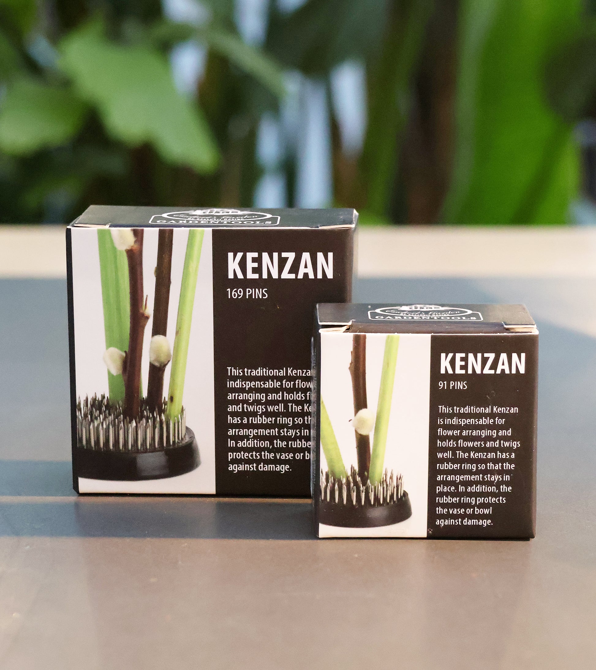 Zwei schwarze Kästen mit der Aufschrift "Ikebana Blumenigel | Kenzan L", jeweils mit Metallspitzen zum Arrangieren von Blumen, mit 169 und 91 Nadeln, stehen auf einem Tisch mit einer verschwommenen grünen Pflanze im Hintergrund.