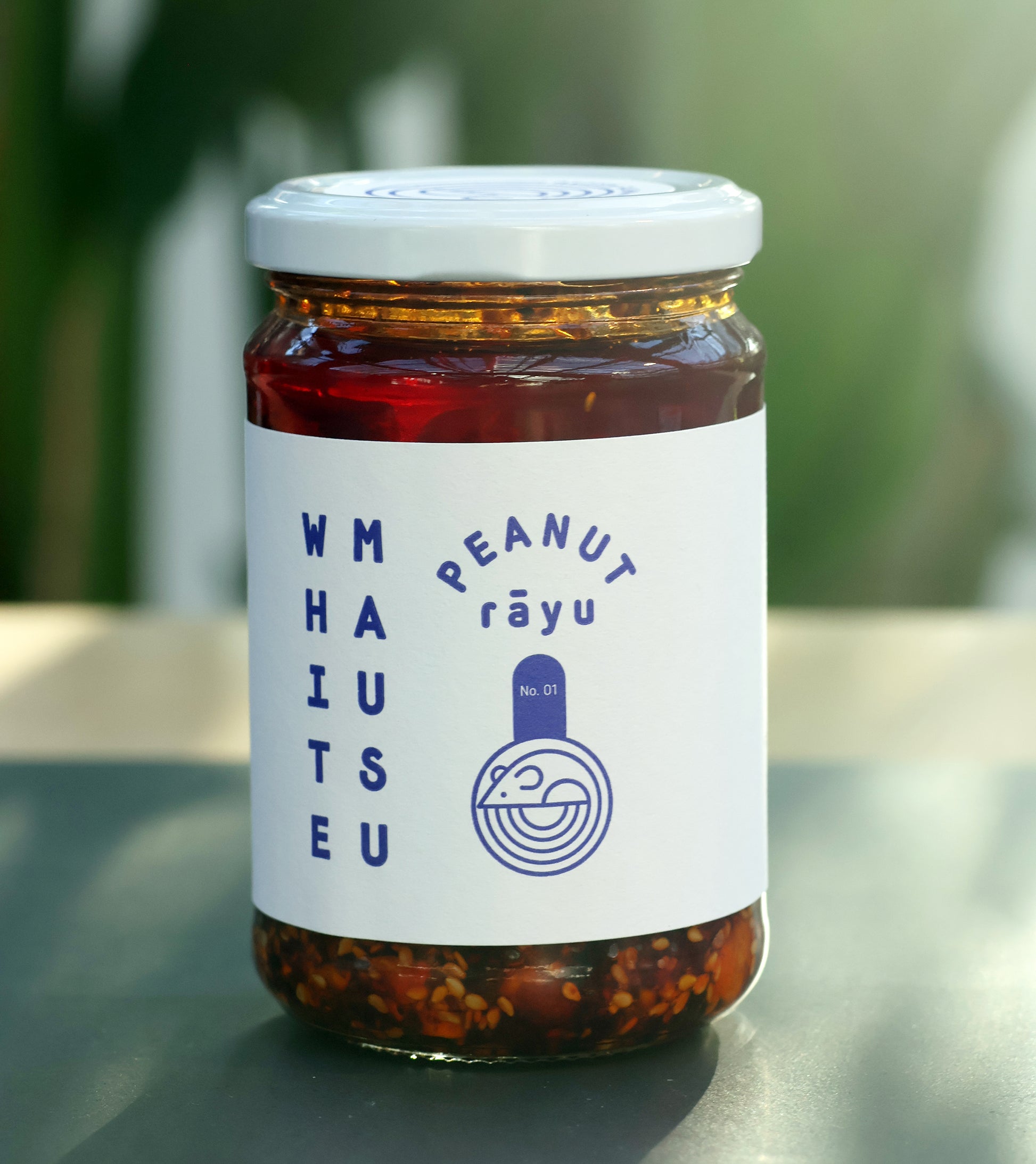 Ein Glas mit der Erdnuss-Rayu-Sauce der White Mausu Ltd. mit weißem Deckel und einem Etikett mit blauem Text und einfachem Logo auf einer grauen Fläche mit verschwommenem grünem Hintergrund.
