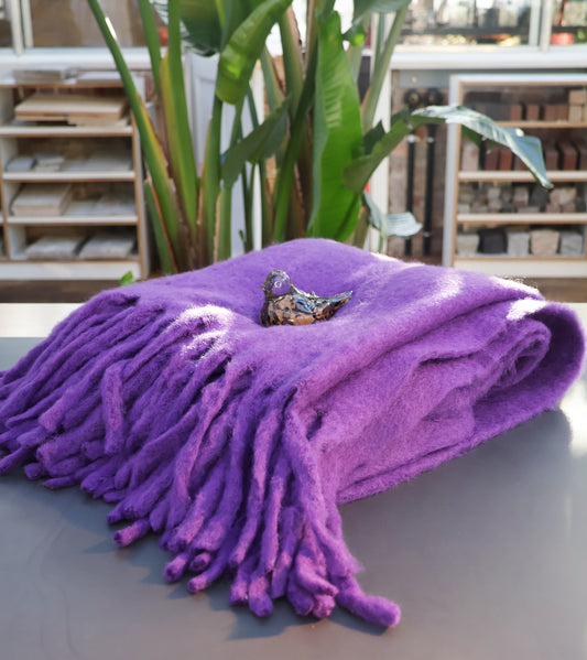 Eine gefaltete Decke aus Wollmischung in lila von Werner Voss mit einer kleinen Keramikvogelfigur darauf ruht auf einer grauen Fläche. Im Hintergrund sind Grünpflanzen und Regale zu sehen.